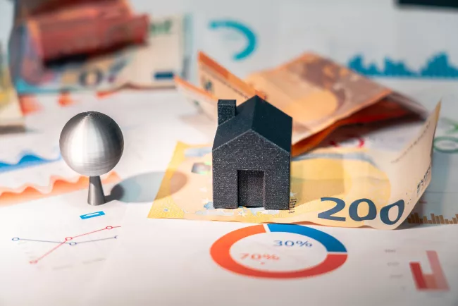Modellino di casa stilizzato di colore scuro, posizionato sopra banconote da 200 euro. Lo sfondo è composto da vari documenti finanziari